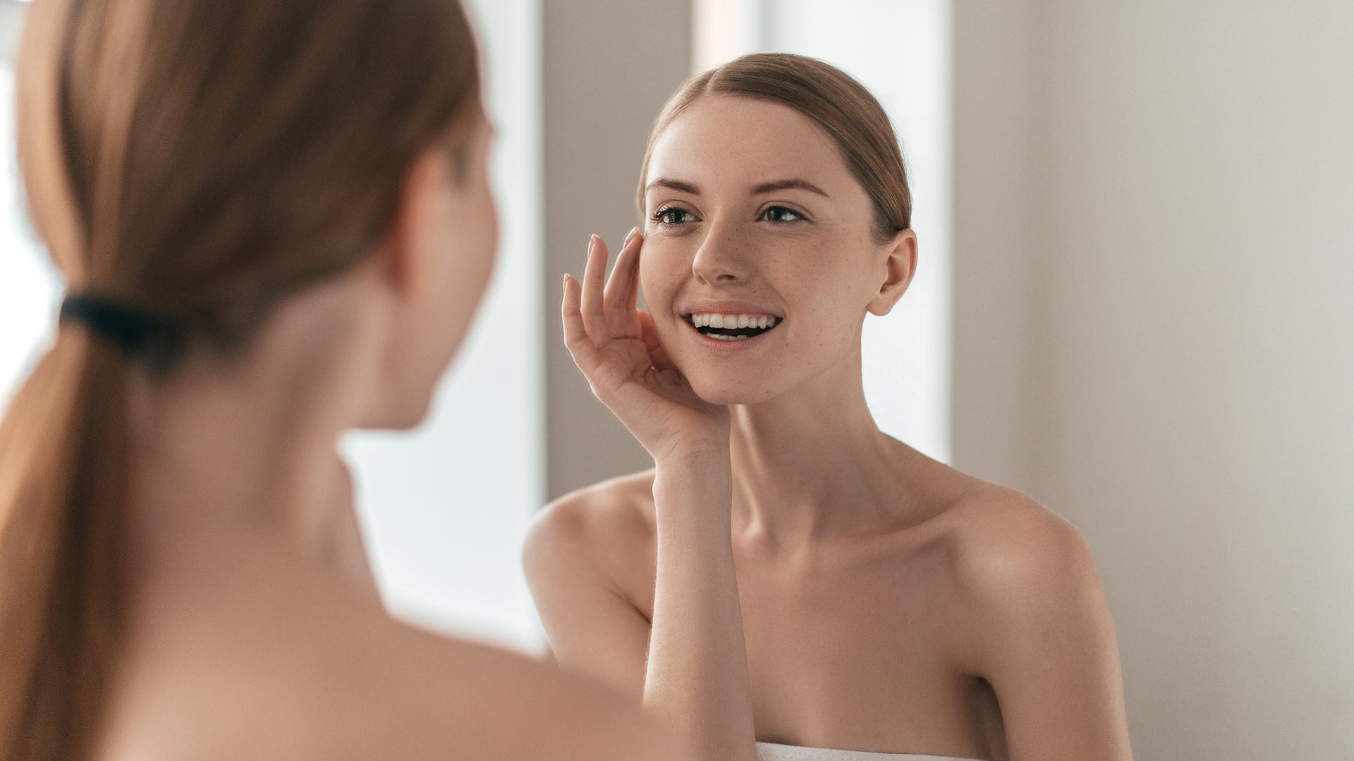 young woman smiling checking clear glowing skin in bathroom mirror